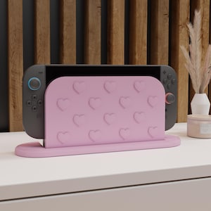 May include: A pink heart-patterned stand for a Nintendo Switch console. The stand is light pink and features embossed hearts. The Switch console is dark grey with blue and red accents. The stand is on a white surface.