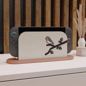 Puede incluir: Una consola Nintendo Switch gris se muestra en un soporte marrón claro. El soporte tiene un panel beige claro con un pájaro posado en una rama. El soporte está sobre una superficie blanca con un fondo de madera.