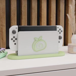 May include: A white Nintendo Switch dock with a light green base and a matching logo. The dock holds the Switch console and two white Joy-Con controllers. The background features a wooden slat wall.