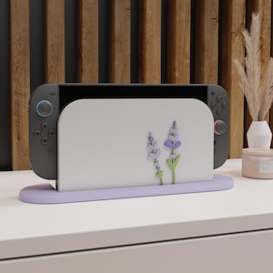 May include: A lilac Nintendo Switch dock featuring a floral design. The dock holds the Switch console, which is black with blue and red controller accents. The design includes purple lavender flowers and green stems.