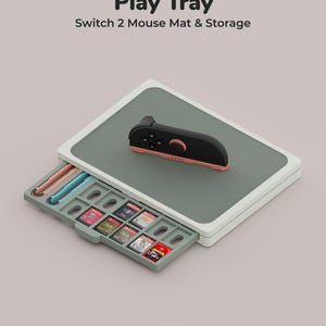 Puede incluir: Una bandeja rectangular Play Tray Switch 2 Mouse Mat & Storage con superficie verde salvia y borde blanco. Un mando de juego negro y melocotón descansa encima. Un cajón deslizante contiene ranuras para cartuchos y dos accesorios.