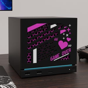 May include: A black computer case with a front panel featuring a pink and white graphic design. The design includes a heart, the text "GG", "XO", and "GAME OVER" over a hexagonal mesh pattern. The base has a thin blue light strip and three ports.