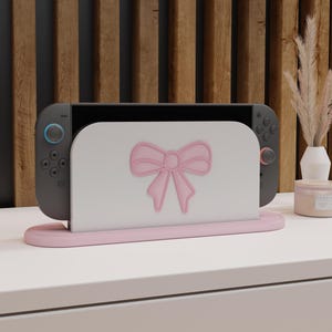 May include: A Nintendo Switch console rests in a pink stand with a white face featuring a pink bow design. The stand and bow are a soft pink color, complementing the console's gray controllers. The background includes a wooden panel and decorative elements.