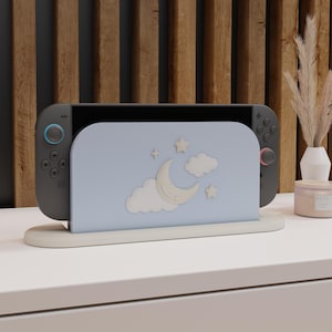 May include: A gray game console with blue and red accents is displayed in a light blue stand. The stand features a crescent moon, stars, and clouds design. The console and stand are on a white surface, with a wooden background.