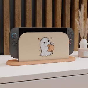Puede incluir: Una consola Nintendo Switch gris con mandos azules y rojos se muestra en un soporte color melocotón. El soporte presenta un fantasma de dibujos animados sosteniendo una calabaza. El fondo incluye un panel de madera y una superficie blanca.