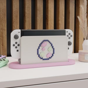 May include: A white Nintendo Switch console with attached white controllers, resting in a pink stand. The console features a pixelated egg design with pink and green accents. The setup is on a white surface.