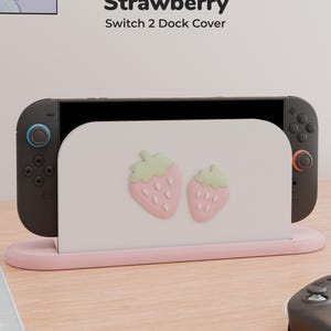 Può includere: Una cover bianca per la docking station di una console da gioco con due icone di fragole in rilievo color rosa chiaro e foglie verde pallido. Il testo "Strawberry Switch 2 Dock Cover" è stampato sopra la console su una base rosa.