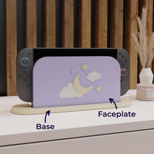 May include: A Nintendo Switch console with a lavender faceplate featuring a crescent moon, stars, and clouds design. The console rests on a beige base. The background includes a wooden wall and decorative items.