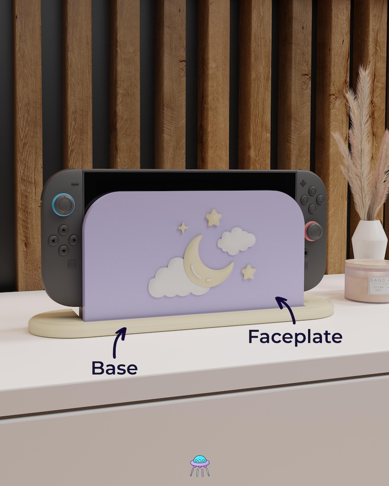 May include: A Nintendo Switch console with a lavender faceplate featuring a crescent moon, stars, and clouds design. The console rests on a beige base. The background includes a wooden wall and decorative items.