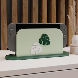 May include: A sage green Nintendo Switch dock with a dark green base and two leaf accents. The dock holds a gray Nintendo Switch console. The background features a wooden panel and a white surface.