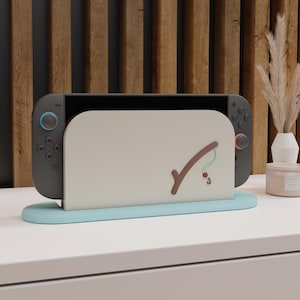 May include: A gray Nintendo Switch console docked in a light blue stand. The stand has a beige back with a brown fishing rod design and a red bobber. The background features a wooden panel.