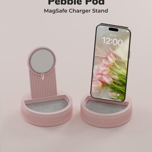 Könnte beinhalten: Zwei hellrosa Pebble Pod MagSafe Ladeständer mit gerippten vertikalen Details und einer kreisförmigen magnetischen Ladefläche. Ein Ständer ist leer, der andere hält ein Smartphone mit einem Blumen-Hintergrundbild und der Uhrzeit 12:00.
