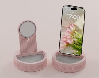 May include: Two light pink Pebble Pod MagSafe charger stands with ribbed vertical detailing and a circular magnetic charging surface. One stand is empty, and the other holds a smartphone displaying a floral wallpaper and the time 12:00.