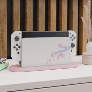 Puede incluir: Una consola Nintendo Switch blanca con Joy-Cons adjuntos, colocada en una base de carga rosa. La base presenta un diseño floral con flores rosas y una rama morada. Fondo con una pared de listones de madera.