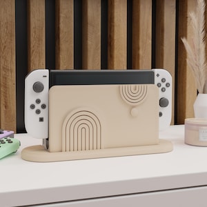 Puede incluir: Un soporte beige para la consola Nintendo Switch con un diseño geométrico. El soporte tiene una ranura para la consola y dos ranuras para los mandos. El soporte está sobre una superficie blanca.