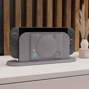 Puede incluir: Un soporte gris para consola diseñado para una Nintendo Switch, con un diseño de consola de juegos retro. El soporte tiene un diseño circular y una base, con detalles en azul y amarillo. Está sobre una superficie blanca.