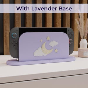 May include: A lavender-colored game console stand with a crescent moon, stars, and cloud design. The stand holds a black game console with blue and red accents. The text "With Lavender Base" is displayed above the stand.