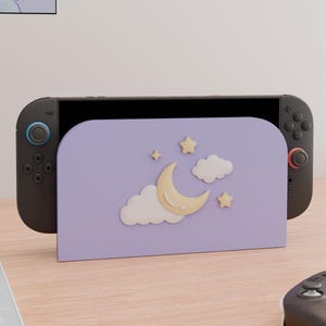 May include: A lavender gaming console dock featuring a raised, cream-colored crescent moon, two white clouds, and two small stars. The dock is attached to a black handheld gaming console with dark gray controllers, one with a blue ring and one with a red ring.