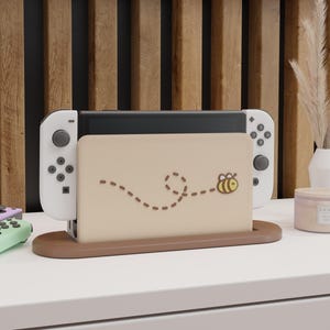 Könnte beinhalten: Eine weiße Nintendo Switch-Konsole mit einem braunen und beigefarbenen Ständer, der mit einem Bienen- und Punktlinienmuster verziert ist. Die Konsole steht auf einer weißen Oberfläche.