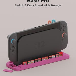 May include: A pink Base Pro Switch 2 Dock Stand with Storage holding a black gaming console and ten game cartridges. The text Base Pro Switch 2 Dock Stand with Storage is printed above the device, and a small UFO icon is at the bottom.