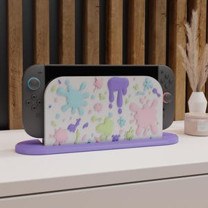 May include: A gaming console dock featuring a white front panel decorated with raised, colorful paint splatters in shades of purple, pink, light blue, and lime green. The dock rests on a solid purple base and holds a dark gray handheld gaming console.