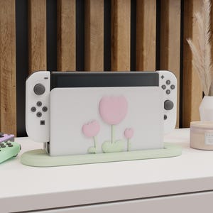 以下が含まれることがあります： ピンクのチューリップと緑の葉が描かれた花柄の白いニンテンドースイッチコンソールスタンド。スタンドは薄い緑色のベースで、スイッチコンソールとジョイコンコントローラー2つを収納するように設計されています。
