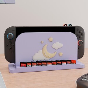 May include: A light purple gaming console dock featuring a crescent moon, two clouds, and two stars on the front panel. The dock includes a row of slots for ten game cartridges and holds a black gaming console with dark gray controllers attached.