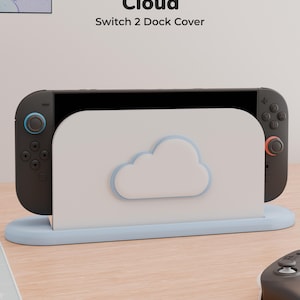 May include: A white and light blue cloud-themed dock cover for a gaming console. The cover features a raised cloud icon on the front panel and is labeled with the text Cloud Switch 2 Dock Cover. The console is docked inside the protective cover.