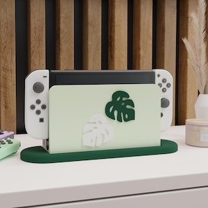 Könnte beinhalten: Ein grüner und weißer Nintendo Switch-Ständer mit einem Ausschnitt von zwei Monstera-Blättern. Der Ständer ist aus Holz und hat eine grüne Basis.