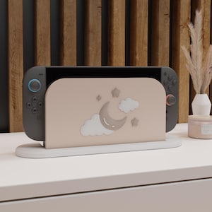 May include: A gray Nintendo Switch console with a light pink stand featuring a crescent moon, stars, and clouds design. The stand is on a white surface, with a wooden slatted background.