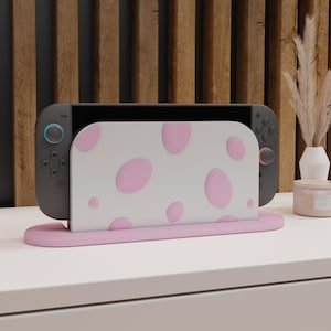 Könnte beinhalten: Eine graue Nintendo Switch-Konsole wird in einem rosa Ständer mit einer weißen Hülle mit rosa Punkten angezeigt. Der Ständer und die Hülle befinden sich auf einer weißen Oberfläche mit einem Holzpaneel-Hintergrund.