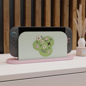 May include: A gray Nintendo Switch console with blue and red controllers is docked in a pink stand. The screen displays a light green design with white flowers, a black cross, and green leaves. The stand sits on a white surface.