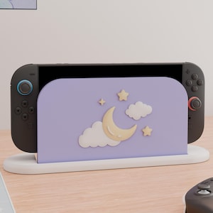 May include: A black handheld gaming console docked in a light purple stand. The stand features a raised, cream-colored crescent moon, two white clouds, and two small stars. The console has black controllers with blue and red accents on the joysticks.