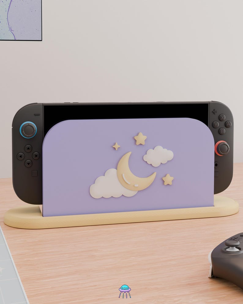 May include: A black handheld gaming console docked in a light purple stand. The stand features a raised, cream-colored crescent moon, two white clouds, and two small stars. The console has black controllers with blue and red accents on the joysticks.