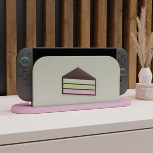 Pode incluir: Um console Nintendo Switch cinza repousa em um suporte rosa. O suporte apresenta uma inserção verde claro com um gráfico de bolo. O fundo inclui uma parede de madeira e uma mesa branca.
