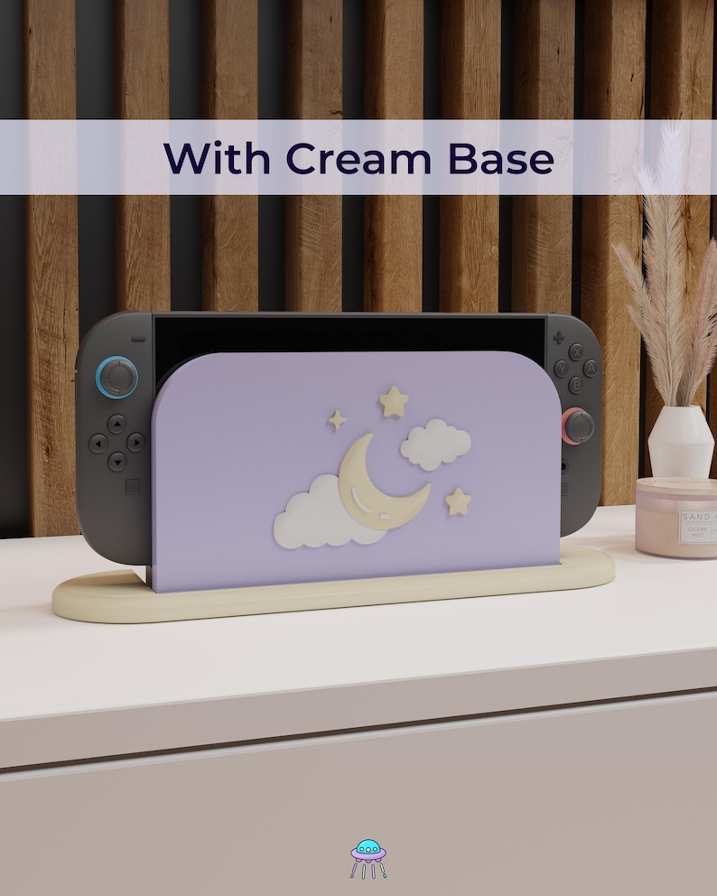 May include: A gray game console with a lavender stand featuring a crescent moon, stars, and clouds design. The stand has a cream base, and the text "With Cream Base" is displayed above the console.