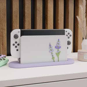 May include: White Nintendo Switch console stand with a lavender floral design. The stand has a purple base and is designed to hold the console upright.