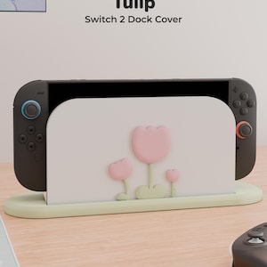 Peut inclure: Une housse de station d'accueil Switch 2 blanche ornée de trois motifs de tulipes roses en relief avec des tiges et des feuilles vert clair, sur une base vert clair. Le texte "Tulip Switch 2 Dock Cover" est inscrit au-dessus de la console noire.