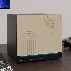 May include: A black rectangular electronic device featuring a beige front panel with embossed concentric arch and circle patterns. The base includes two USB ports, a USB-C port, a small circular power button, and a thin horizontal blue light strip.