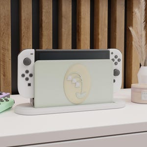Könnte beinhalten: Eine weiße Nintendo Switch-Dockingstation mit einer hellgrünen Frontplatte mit einem Maiglöckchen-Design. Die Dockingstation hält die Switch-Konsole und weiße Joy-Con-Controller. Ein hellgrüner und lila Controller ist im Hintergrund sichtbar.