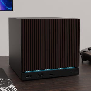 May include: A compact, cube-shaped computer tower featuring a dark brown, vertically slatted front panel. The base is matte black with two USB ports, a central input slot, and a small circular power button, accented by a thin, glowing blue light strip.