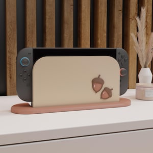 Peut inclure: Une console Nintendo Switch grise est présentée dans un support marron clair avec des motifs de glands. Le support est posé sur une surface blanche, avec un fond en panneaux de bois.
