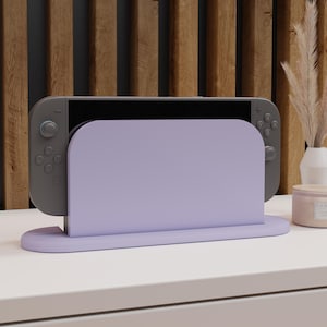 May include: A lilac Nintendo Switch dock with a matching base, holding a grey Switch console. The dock is on a white surface, with a wooden slatted background. A candle and vase with decorative grass are visible.