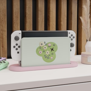May include: A Nintendo Switch console with white Joy-Cons is docked in a pink stand. The console screen features a light green design with white flowers and a black cross. A mint green controller is visible.