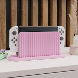Op de afbeelding: Een roze en witte Nintendo Switch console standaard met een geribbeld ontwerp. De console staat in een verticale positie en de standaard heeft een afgeronde basis.