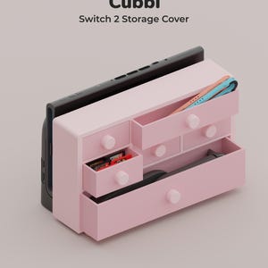 Pode incluir: Uma unidade de armazenamento rosa claro rotulada como Cubbi Switch 2 Storage Cover, com várias gavetas com puxadores redondos. A unidade contém uma consola de jogos preta, cartuchos de jogo e acessórios de comando. O design inclui várias gavetas.