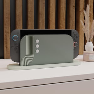 Peut inclure: Une console de jeu grise avec des boutons bleus et rouges, posée dans un support vert sauge. Le support a un design incurvé avec un panneau arrière assorti avec des points blancs et un motif rayé. La console est sur une surface blanche avec un fond en bois.