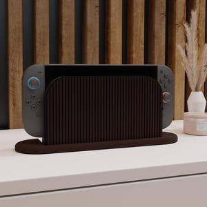 May include: A dark brown wooden gaming console dock featuring a vertical slatted front design. The dock holds a grey handheld gaming console with a black screen, a blue joystick on the left controller, and a red joystick on the right controller.