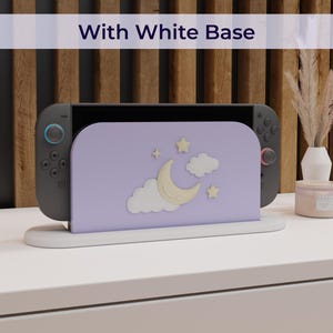May include: A gray game console with a lavender stand featuring a moon, stars, and clouds design. The stand is on a white base, with the text "With White Base" above. The console is on a white surface.