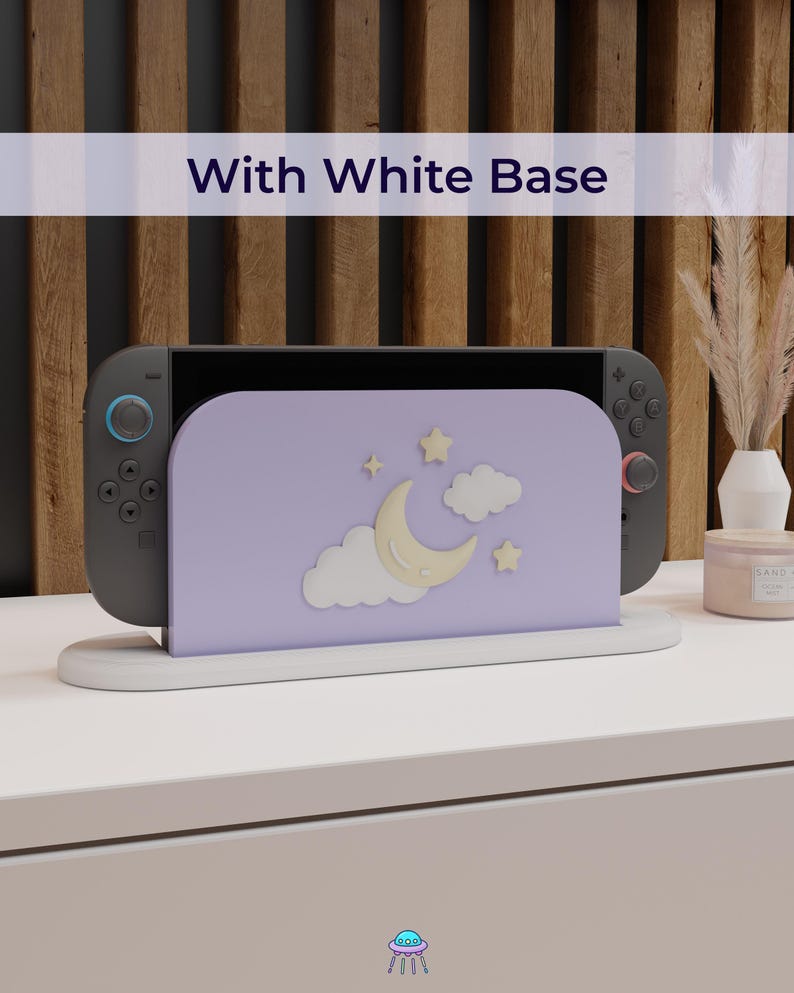 May include: A gray game console with a lavender stand featuring a moon, stars, and clouds design. The stand is on a white base, with the text "With White Base" above. The console is on a white surface.